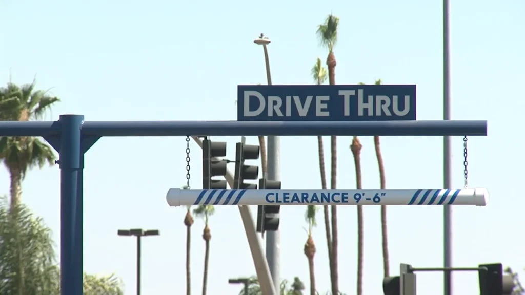 Dutch Bros Locations: 4 A close-up of a blue 'Drive Thru' sign with white text, featuring a clearance bar at 9'6" against a backdrop of palm trees and a clear sky.