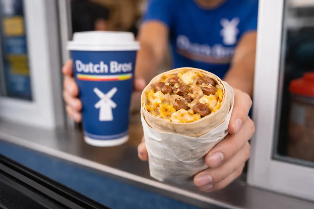 Dutch Bros Breakfast Burrito Price 2 A wrapped breakfast burrito being held at a Dutch Bros drive-thru window with a hot coffee in the background.